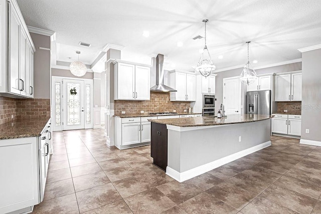 kitchen with wall chimney exhaust hood, stainless steel appliances, white cabinetry, hanging light fixtures, and an island with sink