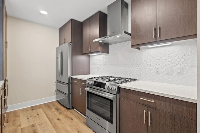 kitchen with wall chimney range hood, light hardwood / wood-style flooring, appliances with stainless steel finishes, tasteful backsplash, and light stone counters