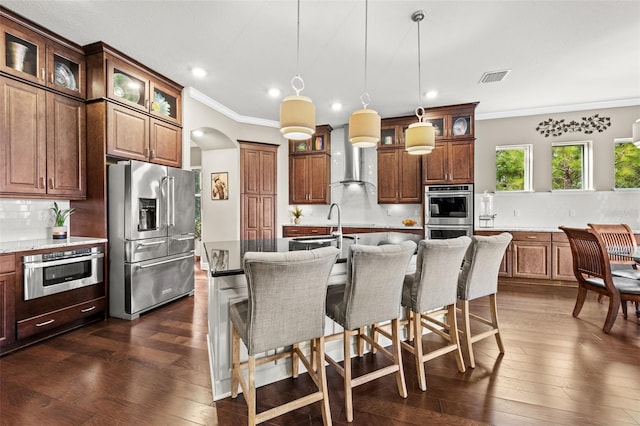 kitchen with a kitchen island with sink, dark wood-type flooring, hanging light fixtures, crown molding, and stainless steel appliances