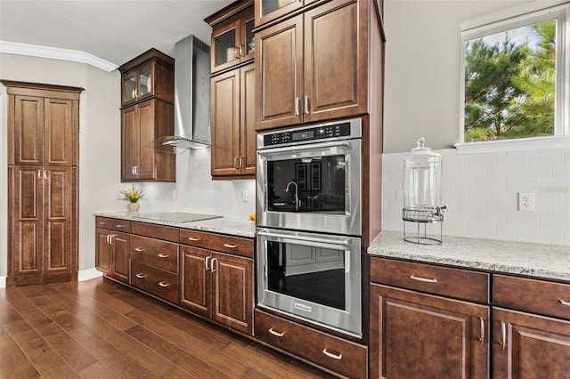 kitchen with light stone countertops, wall chimney exhaust hood, double oven, dark wood-type flooring, and crown molding