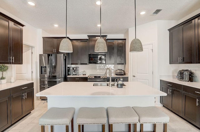 kitchen with dark brown cabinetry, sink, stainless steel refrigerator with ice dispenser, an island with sink, and decorative light fixtures