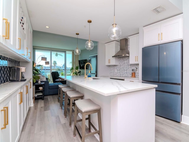 kitchen featuring sink, wall chimney range hood, refrigerator, an island with sink, and white cabinets