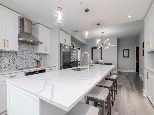 kitchen with stainless steel refrigerator, white cabinets, an island with sink, and wall chimney range hood