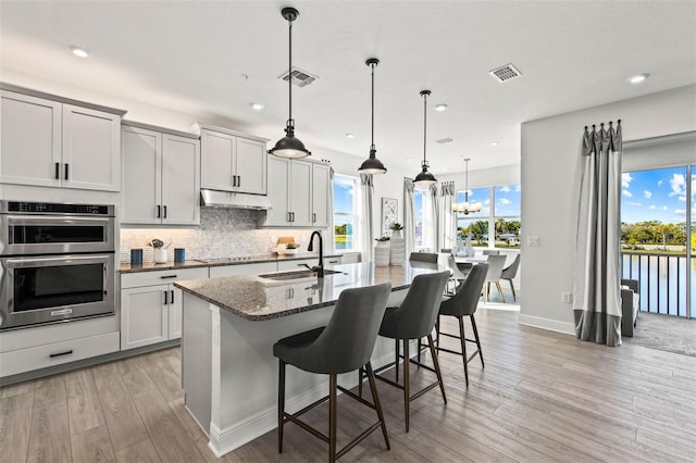 kitchen featuring sink, dark stone countertops, light hardwood / wood-style floors, decorative light fixtures, and a center island with sink