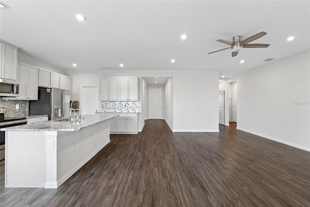 kitchen with light stone countertops, appliances with stainless steel finishes, backsplash, a kitchen island with sink, and dark wood-type flooring