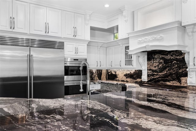 kitchen featuring white cabinets, appliances with stainless steel finishes, decorative backsplash, dark stone countertops, and custom range hood