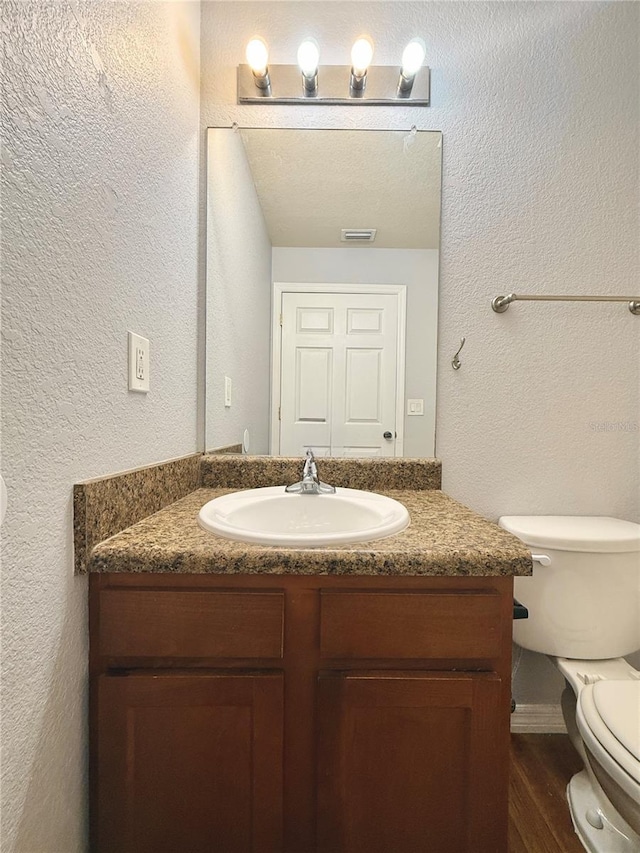 half bathroom with vanity, a textured wall, toilet, visible vents, and wood finished floors