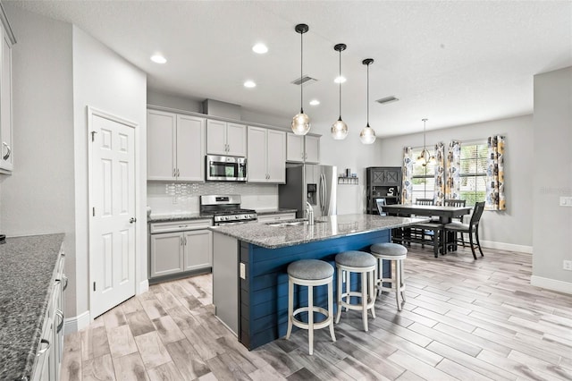 kitchen with dark stone counters, an island with sink, appliances with stainless steel finishes, decorative light fixtures, and light hardwood / wood-style floors