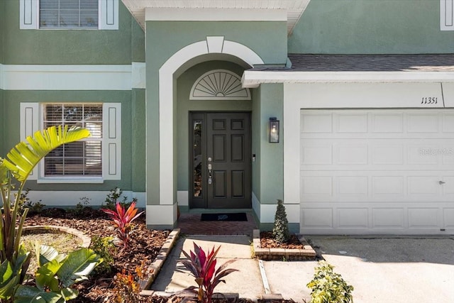 view of exterior entry featuring a garage, roof with shingles, and stucco siding