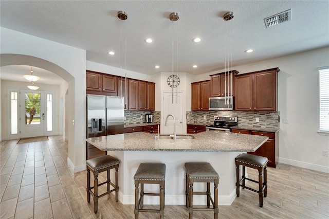 kitchen with light stone countertops, stainless steel appliances, pendant lighting, a breakfast bar area, and a kitchen island with sink