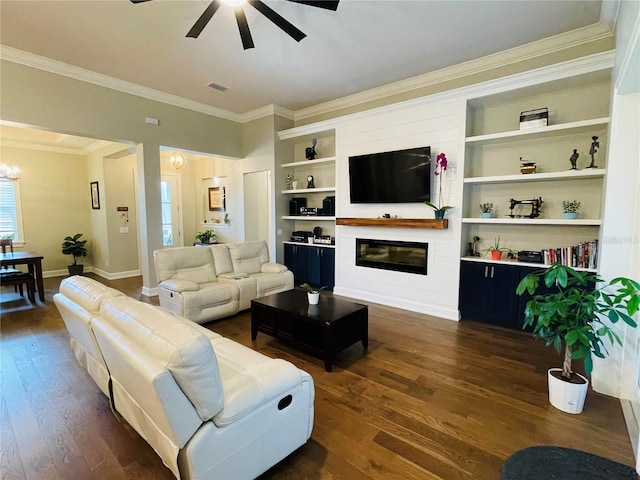 living room featuring a large fireplace, dark wood-type flooring, built in features, ceiling fan with notable chandelier, and ornamental molding