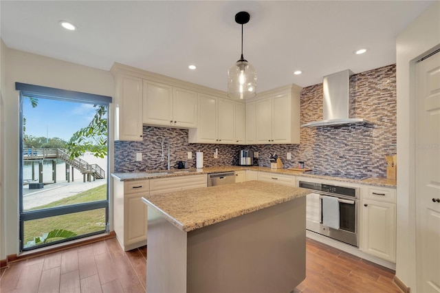 kitchen with wall chimney range hood, sink, hanging light fixtures, appliances with stainless steel finishes, and a kitchen island