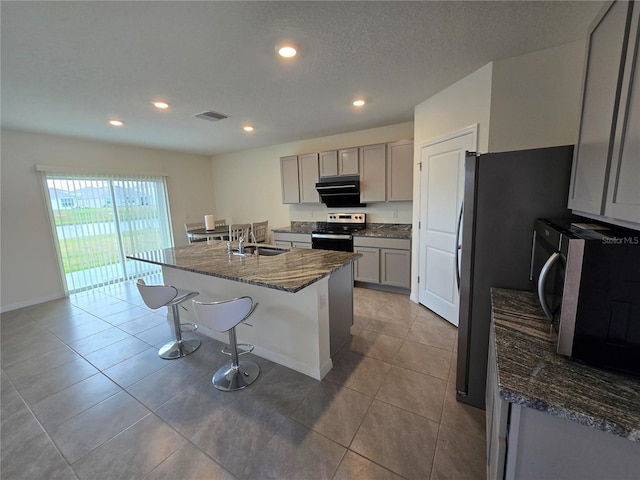 kitchen featuring dark stone counters, a kitchen bar, gray cabinets, a center island with sink, and appliances with stainless steel finishes