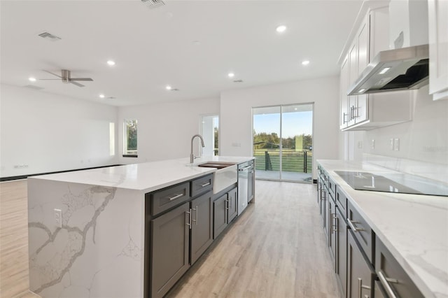 kitchen featuring black electric stovetop, light stone counters, a kitchen island with sink, wall chimney range hood, and white cabinetry