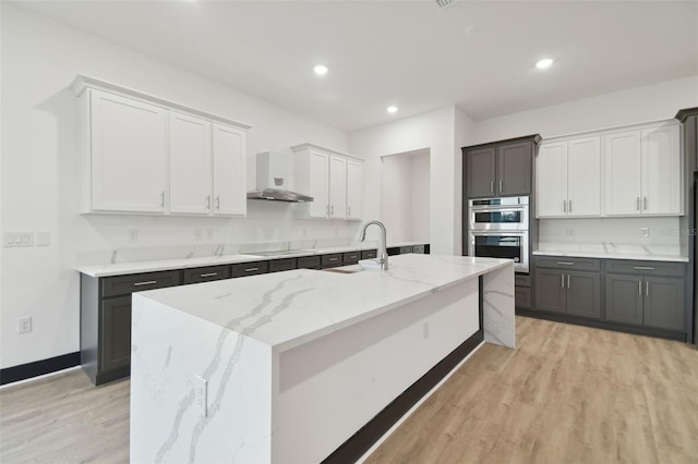 kitchen with light hardwood / wood-style floors, double oven, wall chimney exhaust hood, and a kitchen island with sink