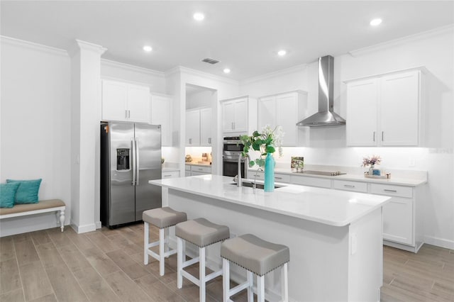 kitchen with wall chimney range hood, white cabinetry, stainless steel appliances, and a kitchen island with sink