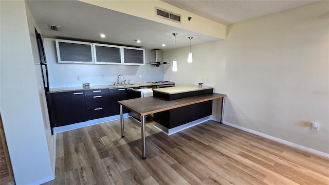 kitchen with kitchen peninsula, sink, wall chimney range hood, wood-type flooring, and pendant lighting