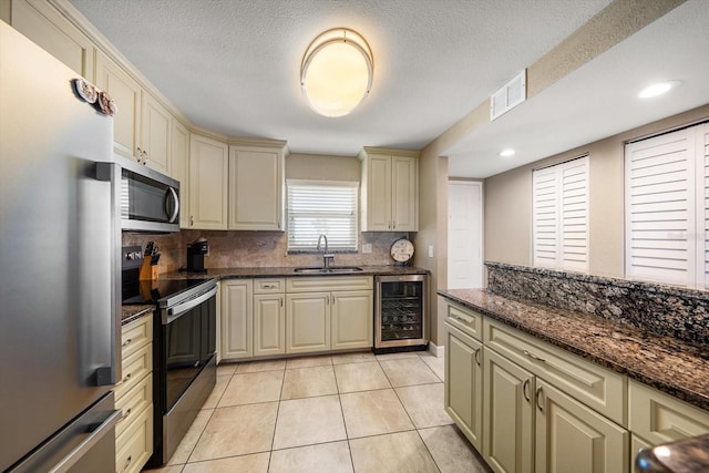 kitchen with dark stone counters, cream cabinets, sink, stainless steel appliances, and beverage cooler