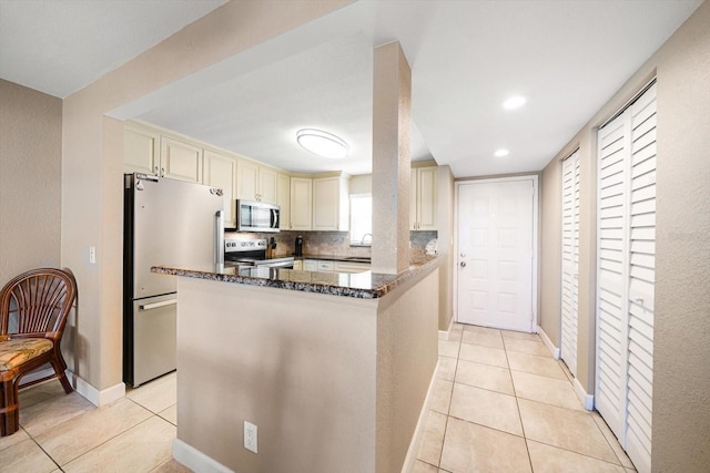 kitchen featuring decorative backsplash, appliances with stainless steel finishes, kitchen peninsula, dark stone counters, and cream cabinetry