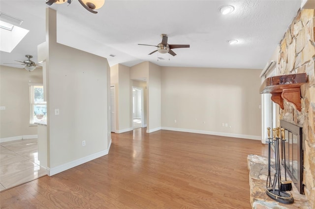 unfurnished living room featuring vaulted ceiling with skylight, a textured ceiling, ceiling fan, light hardwood / wood-style floors, and a stone fireplace