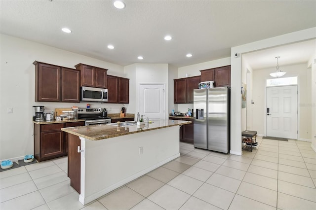 kitchen with light tile patterned floors, a textured ceiling, an island with sink, appliances with stainless steel finishes, and stone countertops