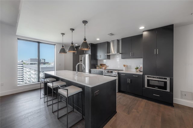 kitchen featuring appliances with stainless steel finishes, a breakfast bar, wall chimney range hood, decorative light fixtures, and a center island with sink