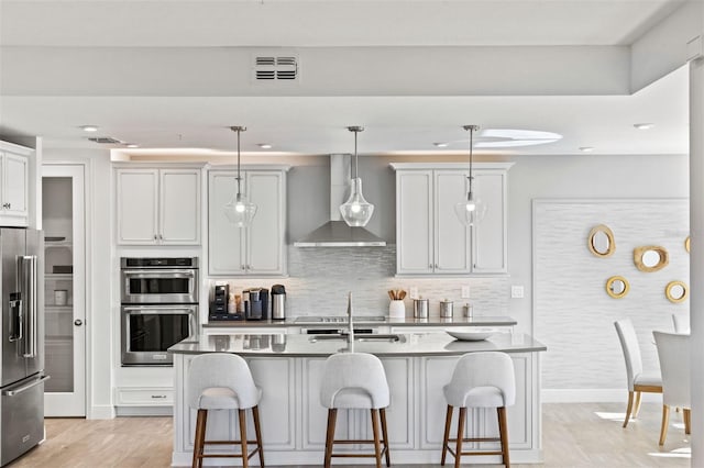 kitchen with appliances with stainless steel finishes, wall chimney exhaust hood, decorative light fixtures, a center island with sink, and a breakfast bar area