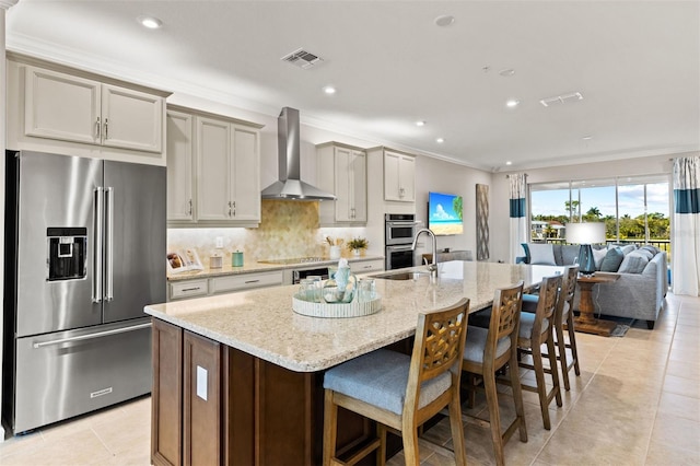 kitchen with wall chimney exhaust hood, a kitchen breakfast bar, light stone counters, an island with sink, and appliances with stainless steel finishes