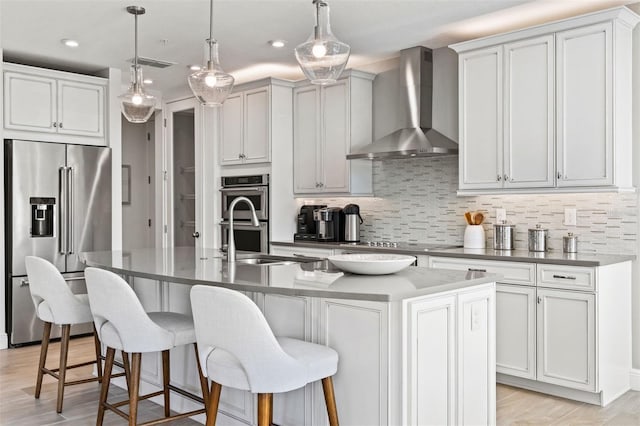 kitchen featuring a kitchen island with sink, white cabinets, wall chimney exhaust hood, appliances with stainless steel finishes, and decorative light fixtures