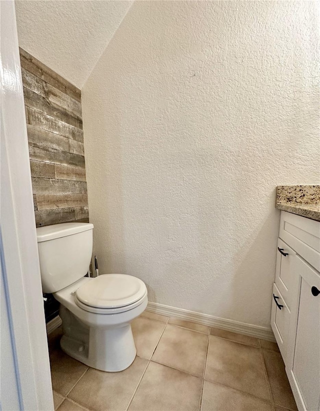 half bathroom with vanity, baseboards, tile patterned flooring, toilet, and a textured wall