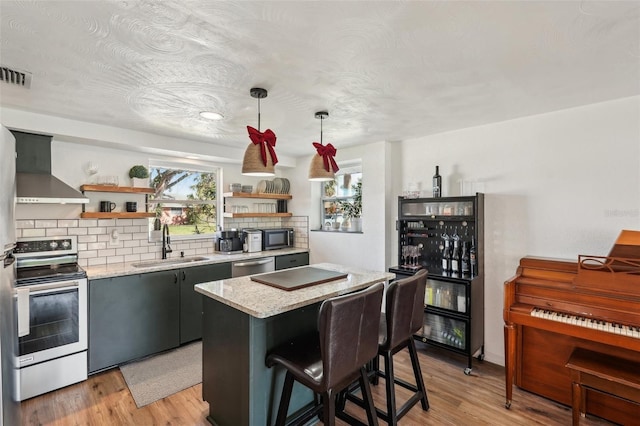 kitchen with sink, wall chimney exhaust hood, hanging light fixtures, light hardwood / wood-style flooring, and appliances with stainless steel finishes