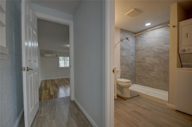 bathroom with a wall mounted air conditioner, toilet, a tile shower, wood-type flooring, and stacked washer / dryer