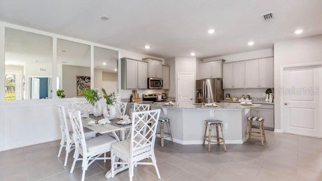 kitchen with gray cabinetry, light stone countertops, an island with sink, appliances with stainless steel finishes, and a kitchen bar