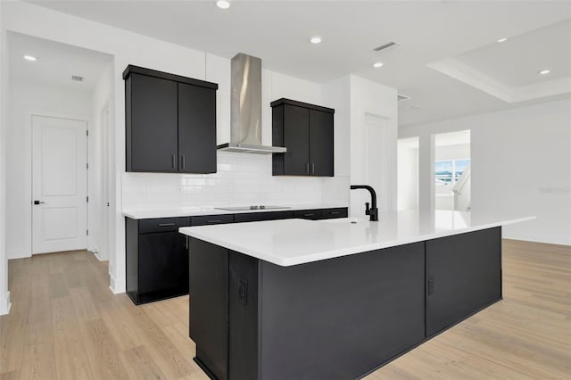 kitchen featuring sink, wall chimney exhaust hood, an island with sink, black electric cooktop, and light wood-type flooring