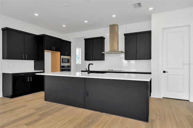 kitchen featuring light hardwood / wood-style floors, wall chimney range hood, a kitchen island with sink, and stainless steel appliances