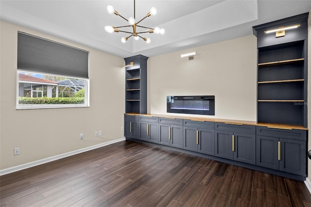 unfurnished living room featuring built in shelves, a tray ceiling, dark wood-style flooring, a notable chandelier, and baseboards