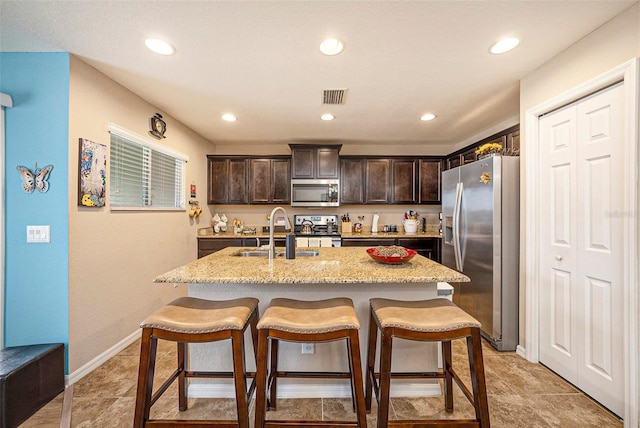 kitchen featuring dark brown cabinetry, sink, light stone counters, a center island with sink, and appliances with stainless steel finishes