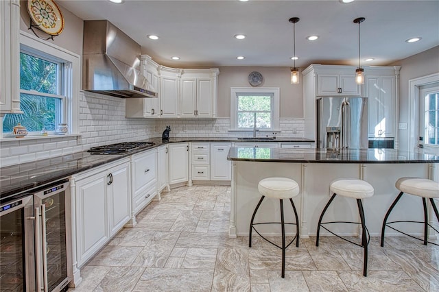 kitchen with white cabinetry, wall chimney exhaust hood, stainless steel appliances, decorative light fixtures, and a kitchen island