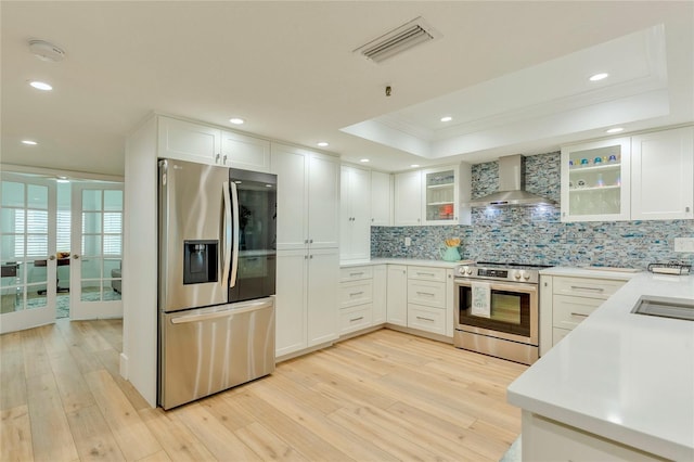 kitchen with stainless steel appliances, a raised ceiling, wall chimney range hood, white cabinets, and light hardwood / wood-style floors