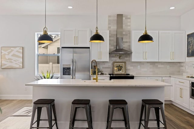 kitchen with a kitchen bar, white cabinetry, appliances with stainless steel finishes, a kitchen island with sink, and wall chimney range hood