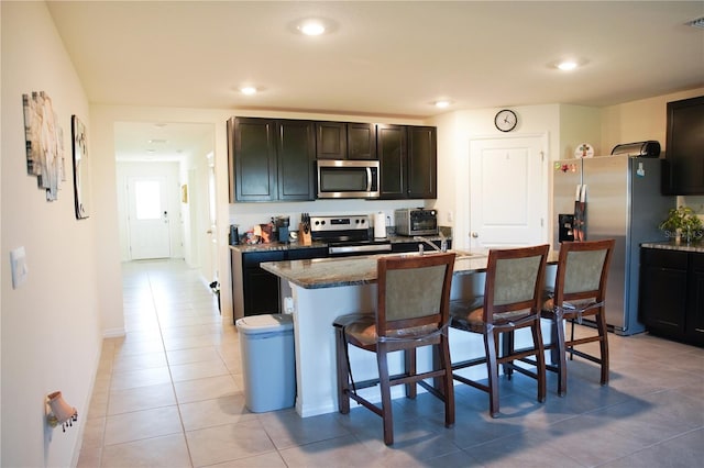 kitchen featuring a kitchen bar, a center island with sink, light tile patterned floors, light stone counters, and stainless steel appliances