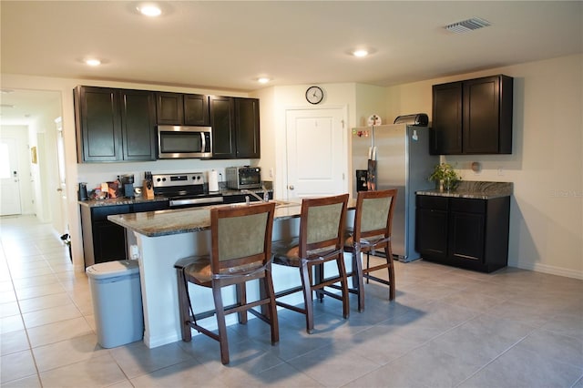 kitchen with a kitchen bar, appliances with stainless steel finishes, light stone counters, a kitchen island with sink, and light tile patterned floors