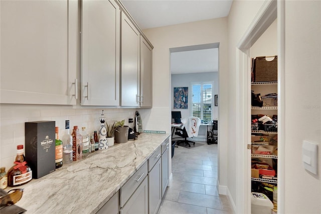 bar with tasteful backsplash, light stone counters, gray cabinetry, built in desk, and light tile patterned flooring