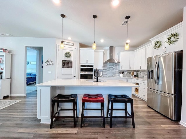 kitchen featuring white cabinets, wall chimney range hood, a kitchen island with sink, and appliances with stainless steel finishes