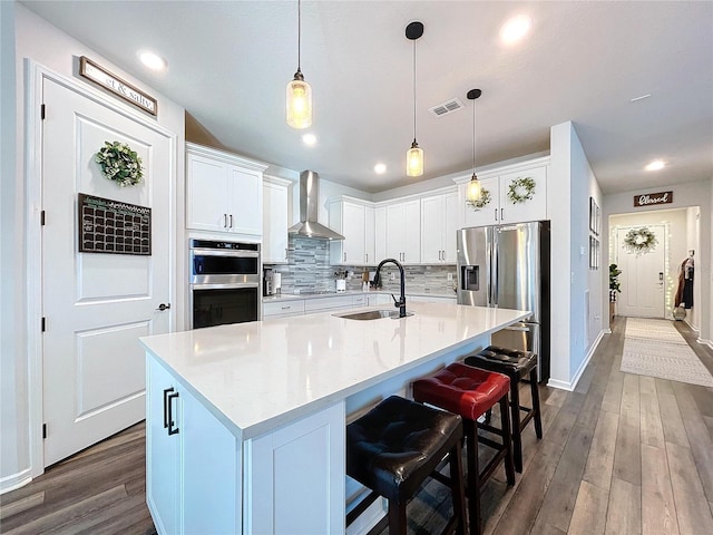 kitchen with white cabinetry, wall chimney exhaust hood, decorative light fixtures, a kitchen island with sink, and appliances with stainless steel finishes