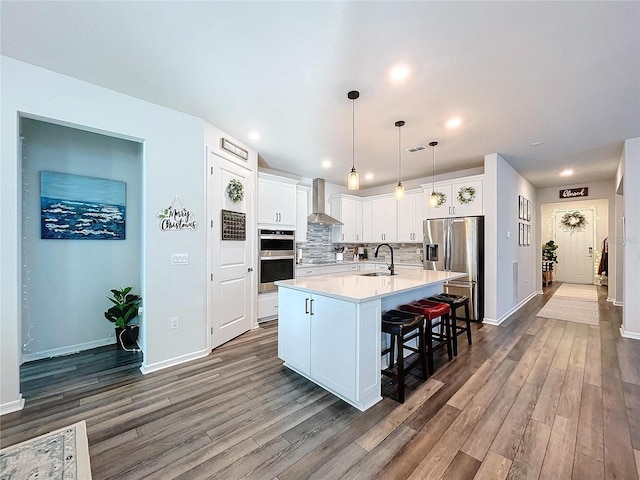 kitchen featuring stainless steel appliances, a kitchen island with sink, wall chimney range hood, sink, and white cabinets