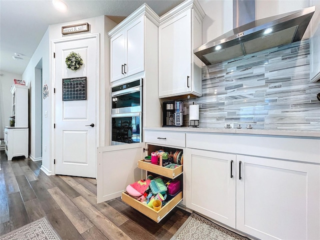 kitchen featuring backsplash, wall chimney range hood, hardwood / wood-style flooring, stainless steel double oven, and white cabinetry