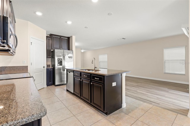 kitchen featuring light stone countertops, dark brown cabinets, stainless steel appliances, a kitchen island with sink, and sink