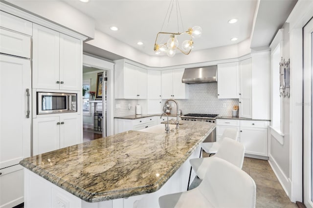 kitchen featuring wall chimney exhaust hood, built in appliances, dark stone countertops, a kitchen island with sink, and backsplash