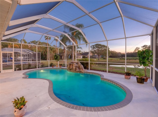 pool at dusk with a patio area, a water view, and glass enclosure